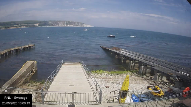 A view of a seaside harbor with a wooden pier extending into the water, surrounded by a metal railing. There are a few small boats floating in the calm blue sea, and white chalk cliffs can be seen in the distance across the bay. The sky is partly cloudy with patches of blue. In the foreground, there are some yellow safety equipment, including a life ring and a cone, along with a small yellow inflatable boat and a blue container. The scene is captured from a webcam and includes a timestamp showing the date as June 20, 2025, at 1:00 PM, with the label "RNLI - Swanage."