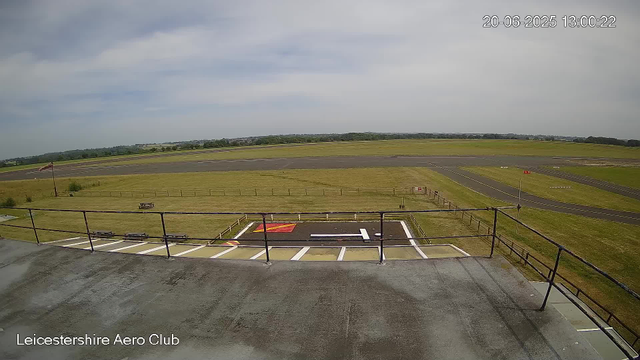 A view from a webcam at Leicestershire Aero Club showing an empty airfield with a runway extending into the distance under a partly cloudy sky. In the foreground, there is a small helipad marked with a white 'H' and red and yellow markings. A railing surrounds the viewing area, and a windsock is visible on the left side. The timestamp on the image reads 20-06-2025 13:00:22.