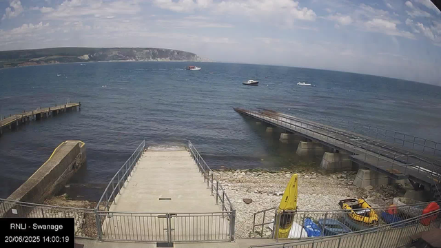 A view of a seaside scene featuring a concrete pier extending into the water, with a gentle shoreline composed of rocks and pebbles. To the right, there is a small area with safety equipment, including yellow lifeguard gear and red equipment. Several boats are floating on the calm sea, and a larger boat is seen moving across the water. In the background, there are green hills and cliffs under a partly cloudy sky.
