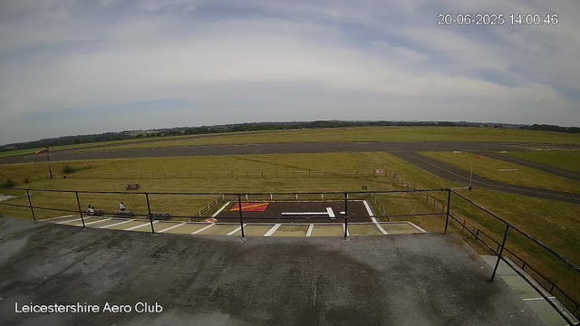 An empty aircraft parking area with a small helicopter pad in the foreground, two people sitting on a bench near the pad, a windsock on a pole to the left, and a wide view of a runway and open grassy fields stretching into the distance under a partly cloudy sky. The image is from the Leicestershire Aero Club, with a timestamp of June 20, 2025, at 14:00:46.
