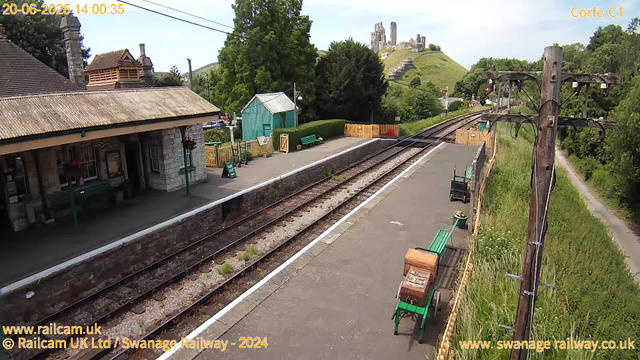 A small train station platform with a single set of railway tracks running through it. There is a green bench and a wooden luggage box on the platform, along with some potted flowers. A building with a tiled roof and hanging flower baskets is on the left side. A small blue-green shed is behind the platform. On the right, there are two wooden utility poles with attached cables, and a narrow dirt path runs alongside the station to the right. In the background, there is lush green foliage, a hill with historic ruins or castle remains at the top, and a gentle sky with light clouds. The date and time displayed on the image are June 20, 2025, at 14:00:35.