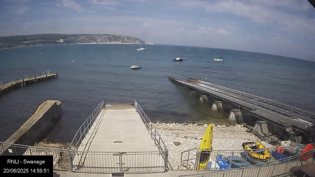 A daytime view of a seaside dock area with small boats anchored in the water. There is a concrete platform with metal railings leading into the water on the left side, and a larger floating platform on the right connected to another structure. The shoreline has rocky terrain, and a few boats and small watercraft are visible. In the background, cliffs and a distant coastline can be seen under a partly cloudy sky.