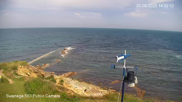 A view of the coastline from a webcam, showing the sea with gentle waves, rocks along the shore, and a weather station with anemometers and other instruments in the foreground. The sky is partly cloudy, and the timestamp in the upper right corner reads "20-06-2025 14:59:52".