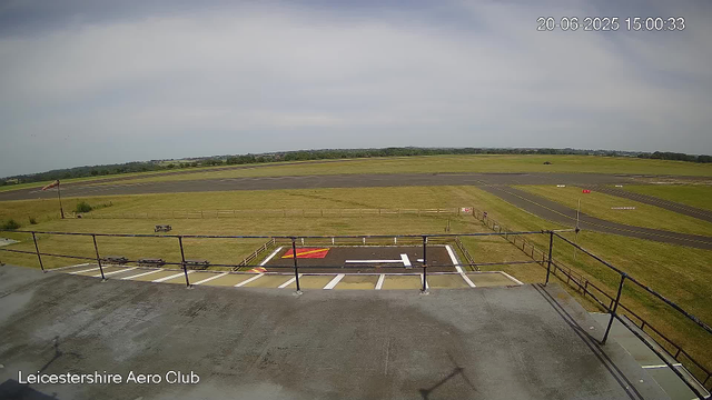 A view from a webcam at Leicestershire Aero Club showing an empty helipad and surrounding airfield. The sky is partly cloudy, and there is an open grassy field beyond the airfield with a few distant trees. A weathercock and some small structures are visible, and the area appears quiet with no aircraft or people present. The timestamp on the image reads 20-06-2025 15:00:33.