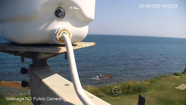 A view from a webcam showing the ocean horizon with clear blue sky. In the foreground, there is a mounted weather or surveillance device attached to a metal stand, with a white cable connected to it. Below, there is a grassy area with a few small bushes and flowers, and some rocks near the water. The timestamp reads "20-06-2025 16:00:28," and the camera label indicates it is from the "Swanage NCI Public Camera."