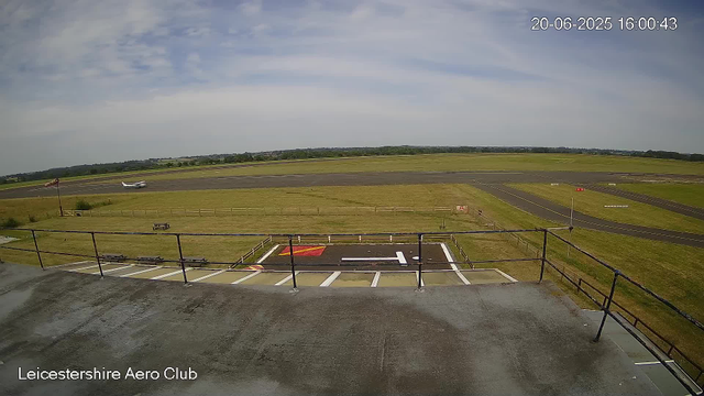 A view of LeicesterShire Aero Club's airfield with a small airplane taxiing on the runway in the distance. The foreground shows a rooftop and safety railing, with a parking space marked with a large white 'H' for helipad. The sky is partly cloudy with patches of blue, and the scene appears to be taken during daytime.