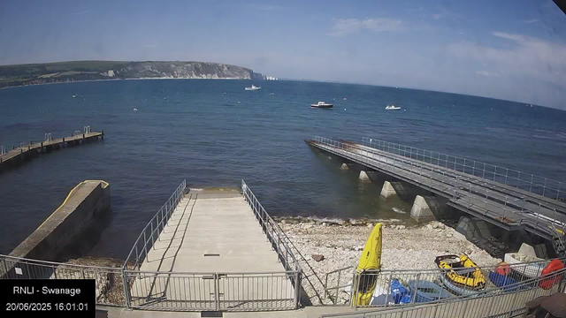 A view of a seaside area with calm blue water, a pier extending into the water, and a boat in the distance. In the background, white cliffs rise along the coast under a partly cloudy sky. At the bottom of the image, there are small boats or kayaks, a yellow life jacket, and some equipment on the rocky shore, all behind a metal fence. The image is labeled "RNLI - Swannage" and timestamped "20/06/2025 16:01:01."
