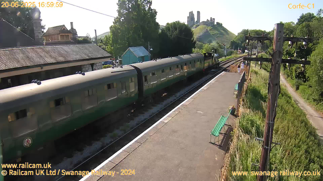 A green passenger train is stopped at a small railway station platform, with a few people standing on the right side near the edge. The platform is bordered by a low fence with some potted plants and a green bench. To the right, a tall wooden utility pole with supporting wires extends upward, and a narrow dirt path runs alongside it. In the background, there are houses with pitched roofs, a small blue shed, lush green trees, and a hillside with ruins or old structures at the top. The sky is clear and sunny, and the timestamp shows the date as June 20, 2025, at 4:59 PM.