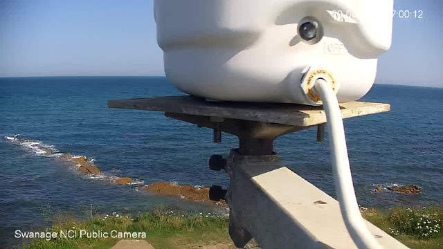 A close-up view of a seismometer instrument mounted on a wooden platform overlooking the ocean. The instrument is white with a small camera and is connected by a white cable. The background shows the blue sea with gentle waves and some rocks near the shoreline, under a clear sky. The text "Swanage NCI Public Camera" is visible in the bottom left corner.