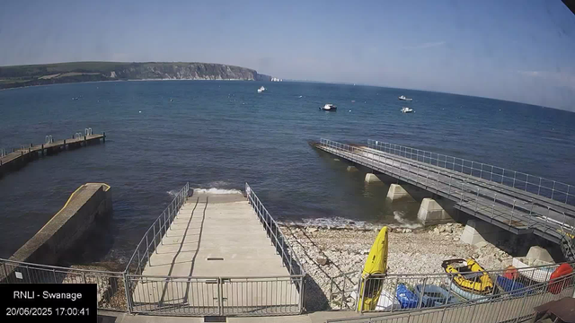 A view of the seaside at Swannage on a sunny day, with boats floating on the water. In the foreground, there is a small concrete pier extending into the sea, flanked by railings. To the right, there's a raised walkway with railings, supported by concrete pillars. On the pebbled beach area below, a yellow surfboard and a yellow pedal boat are visible, along with some parked cars and equipment. The ocean stretches out with a distant shoreline and cliffs visible on the horizon. The sky is partly cloudy with patches of blue.