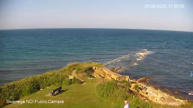 A scenic view of a coastal area with a grassy promenade, a bench, and some greenery in the foreground. The ocean stretches out to the horizon with gentle waves. The sky is clear with no visible clouds. There is a timestamp in the upper right corner showing the date and time as June 20, 2025, at 5:59:56 PM. A person with light hair and a light-colored top is standing near the bottom right corner. The bottom left corner has the text "Swanage NCI Public Camera."