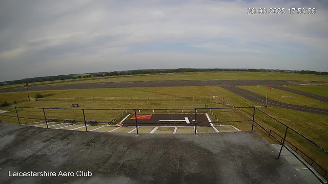 A view from a webcam at Leicestershire Aero Club shows an empty helicopter landing pad in the foreground with white markings, a black surface, and a red and white hazard sign. Beyond the pad, there is a small grassy area enclosed by a wooden fence, and further in the distance, a paved taxiway and runway with markings. The sky is partly cloudy with patches of blue visible, and the scene is well-lit. In the upper right corner, there is a timestamp indicating the date and time as 20-06-2025 17:59:56. The lower left corner displays the text "Leicestershire Aero Club".