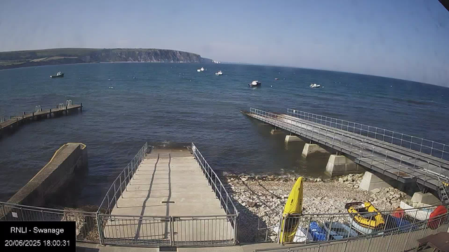 A view of a harbor area with a concrete pier extending into the water. There are several boats floating on the sea, and a distant landmass with cliffs is visible across the water. In the foreground, a fenced area contains a yellow kayak and a yellow vehicle. The sky is mostly clear with some light clouds. The timestamp shows the date as June 20, 2025, at 6:00 PM.