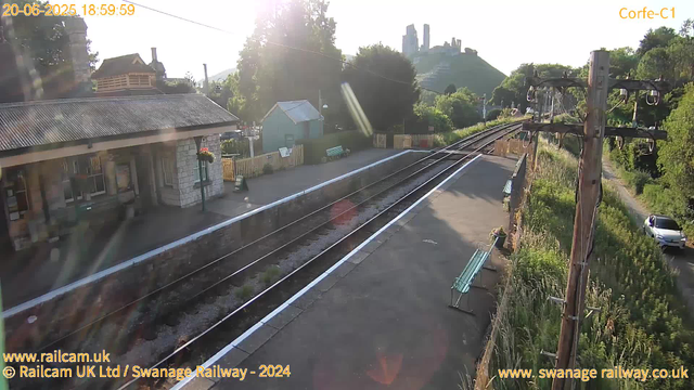 A railway station platform is shown with railway tracks running parallel. To the left, there is a small stone and brick station building with a dark tiled roof, flower baskets hanging outside, and a sign that says "Way Out." On the platform, there are green benches and flower pots, with a white line marking the edge next to the tracks. To the right, there is a tall wooden electricity pole with multiple wires and insulators. In the background, lush green trees, parking area with a white car, and a hillside with castle ruins or old structures are visible. The image is sunlit, with the sun low in the sky, creating lens flares and a warm glow. The timestamp indicates the date and time as June 20, 2025, at 18:59:59.