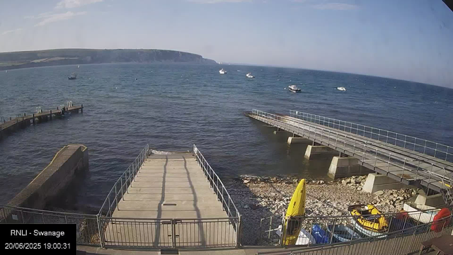 A view of a seaside location with a concrete pier extending into the water, accompanied by a smaller dock and boat ramp nearby. Several boats are floating on the calm ocean. In the foreground, there are yellow rescue boats and equipment, and a fenced area with parking underneath a clear blue sky. In the distance, there are cliffs or hills along the coastline.