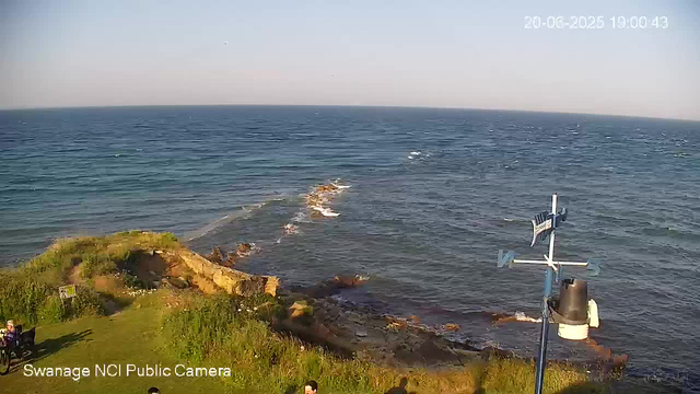 A view of the coastline with a grassy area in the foreground, a rocky shoreline, and the ocean extending to the horizon. There is a weather vane and anemometer mounted on a pole near the water's edge, with some people visible near the grassy area. The sky is clear, and the photo is timestamped "20-06-2025 15:00:43" in the top right corner.