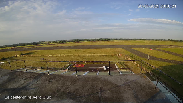 A view from a webcam shows an airfield with grassy fields and runways extending into the distance, under a partly cloudy sky. In the foreground, there is a helipad painted with a white "H" and a red square, surrounded by a metal railing. The timestamp in the top right corner reads "20-06-2025 20:00:24," and the bottom left corner displays "Leicestershire Aero Club."