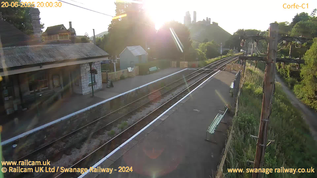 A view of a small railway station platform at sunset, with the sun low in the sky and shining over the horizon. There are empty benches along the platform, a small stone building with windows, and a wooden fence separating the platform from the tracks. Overhead electric lines run alongside the tracks, supported by a metal pole on the right. In the background, there are trees, a hill, and some buildings with chimneys. The image is bright, with lens flare and sunlight glare visible. The date and time are displayed at the top, along with some web and copyright information at the bottom.
