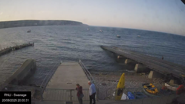 A view of a seaside promenade at dusk with a pier extending into the water. Several boats are visible on the calm sea, and a large cliff stretches along the horizon. Two people are walking together near the lower part of the image, and there is a yellow caution sign and some equipment (such as a small yellow inflatable boat) on the right side. The image is labeled "RNLI - Swanage" with a timestamp of 20/06/2025 20:00:31.