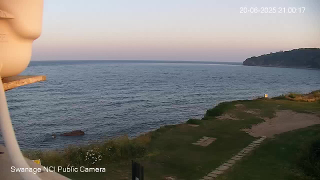 A view of the ocean at sunset with a distant hill on the right side. The sky is clear with a gradient from light to darker blue. In the foreground, there is a grassy area with a paved stone path. The timestamp in the top right corner shows "20-06-2025 21:00:17," and the bottom left indicates "Swanage NCI Public Camera."