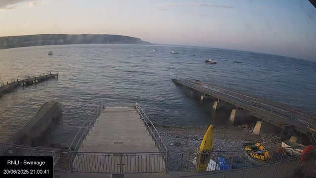 A view of a harbor with a concrete pier extending into the water, surrounded by a metal railing. Several boats are anchored in the distance on a calm sea. In the foreground, there is a yellow caution sign and some equipment or bags placed near the railing. The background features a low, rolling coastline with cliffs under a sky transitioning from dusk to night.