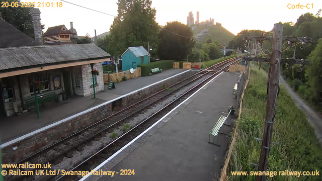 A small railway station platform during sunset, with empty benches, a green hut, a sign reading "WAY OUT," and a building with a tiled roof and chimney on the left. Overhead electric lines run along the tracks, which extend into the distance. In the background, there is a hill with a castle or historic ruin atop, surrounded by trees and greenery. To the right, a narrow path runs parallel to the tracks, bordered by tall grass and trees. The scene is calm and quiet, with a warm glow from the setting sun.