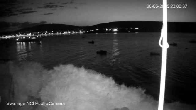 A black-and-white photo taken from a webcam at night showing a body of water with gentle waves. Several small boats are floating on the water. In the background, there are hills with scattered lights, possibly from buildings or houses. The sky has some clouds. The timestamp in the upper right corner reads "20-06-2023 23:00:37," and the bottom left corner indicates "Swanage NCI Public Camera."