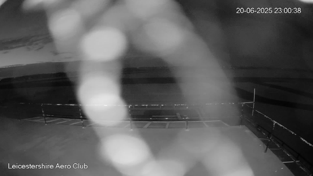 A black-and-white image from a webcam shows an empty outdoor sports field at night, with the name "Leicestershire Aero Club" at the bottom. The timestamp in the top right corner indicates the date and time as June 20, 2025, at 11:00 PM. Large blurry white circles are scattered in the foreground. The field is surrounded by a low fence and is illuminated faintly by distant lights. The sky appears dark and clear.