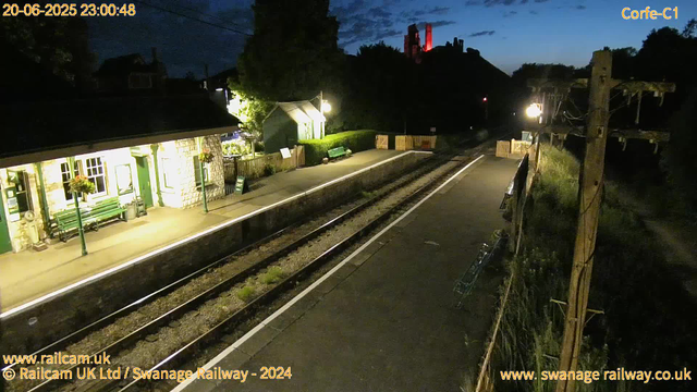 A small railway station platform at night, illuminated by a few street lamps. On the left, there is a stone building with green doors and windows, decorated with hanging flower baskets, and a green bench outside. The platform is empty, with a sign and some benches. Train tracks run parallel to the platform, stretching into the darkness. In the background, atop a hill, a castle-like structure with red lighting is visible. A wooden fence and a utility pole with wires are on the right side of the platform. The sky is dark, with some clouds visible. The timestamp indicates the photo was taken at 23:00 on June 20, 2025.
