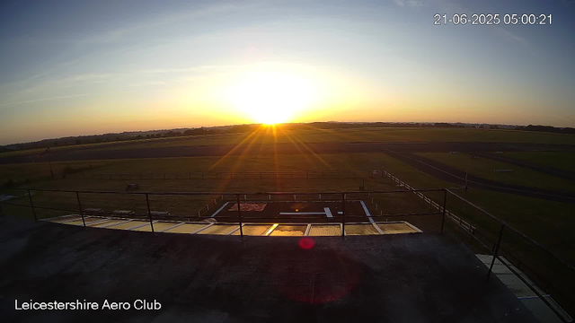 A sunrise over an open grass field at Leicestershire Aero Club, with the sun just above the horizon casting a warm golden light. The sky is clear with faint streaks of clouds, and the landscape appears calm and expansive. There is a balcony or platform in the foreground with a railing, and some fencing and runway markings can be seen in the distance. The timestamp shows the scene is early morning on June 21, 2025.
