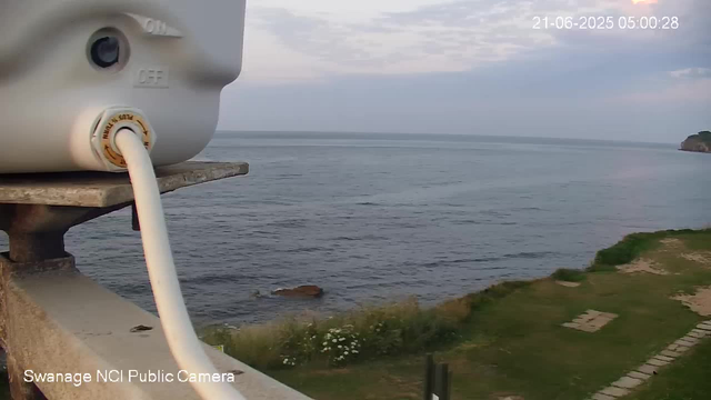 A view of the ocean with a rocky shoreline and some greenery, taken from a webcam mounted on a pole. A small part of a weather sensor with a cable is visible in the foreground on the left side of the image. The sky is partly cloudy and the water appears calm. A timestamp in the top right corner shows "21-06-2025 05:00:28."
