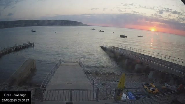 A scenic view of a harbor at sunrise shows several boats floating on calm water, with a distant horizon and a landmass on the left. The sky is partly cloudy with the sun just beginning to rise, casting a warm glow on the water. In the foreground, there is a small dock and a promenade with railings, along with some equipment and vehicles nearby. A sign on the bottom left indicates the location as "RNLI – Swannage" and the date and time as "21/06/2025 05:00:23."