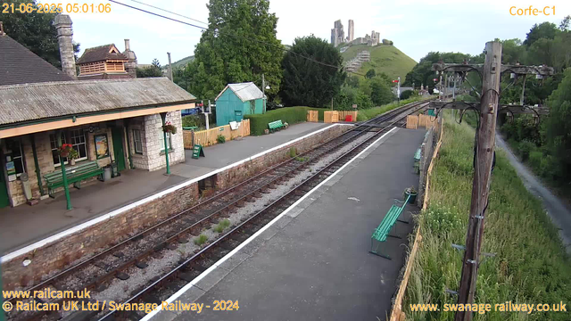 A small train station platform with green benches, a stone building with flower baskets hanging from the roof, and a green shed in the background. The railway tracks run through the middle of the image, curving slightly into the distance where a castle ruins sit atop a grassy hill. There are overhead electrical lines supported by wooden poles along the right side of the tracks. The time and date are displayed in the top left corner as "21-06-2025 05:01:06." The scene appears peaceful and quiet with lush greenery surrounding the station.