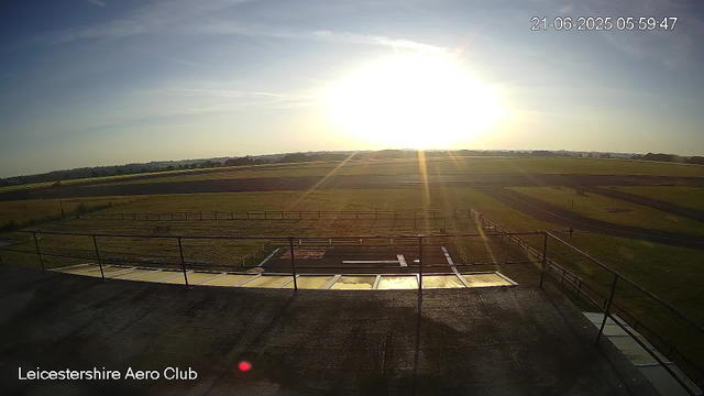 The image shows a landscape at sunrise, with the sun low on the horizon illuminating the sky with bright light. In the foreground, there is a flat rooftop with safety railings. Beyond the rooftop, there are multiple grassy fields and a running track or similar oval-shaped pathway, likely part of a sports facility. The background features a distant horizon with trees and open land under a mostly clear sky with a few wispy clouds. The date and time are displayed in the top right corner: June 21, 2025, at 05:59:47. The text "Leicestershire Aero Club" is visible in the bottom left corner.