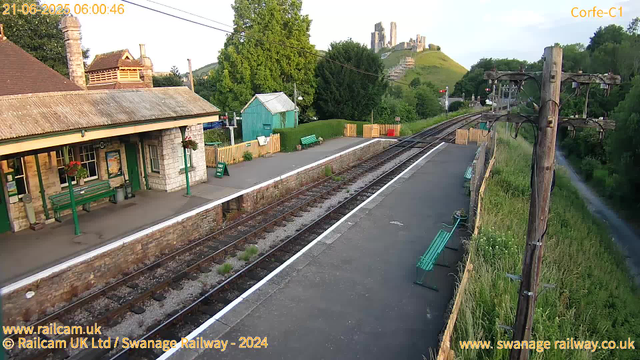 A small rural train station platform with a building on the left featuring a stone facade, green benches, hanging flower baskets, and a "WAY OUT" sign. There are wooden fences and benches along the platform. Overhead, there are electrical wires for the trains. A railway track runs alongside the platform, extending into the distance. In the background, on a green hillside, there is a medieval castle ruin. The scene is captured in early morning light, with the timestamp showing 06:00:46 on June 21, 2025. The station is identified as Corfe-C1, and the image is credited to Swanage Railway, 2024.