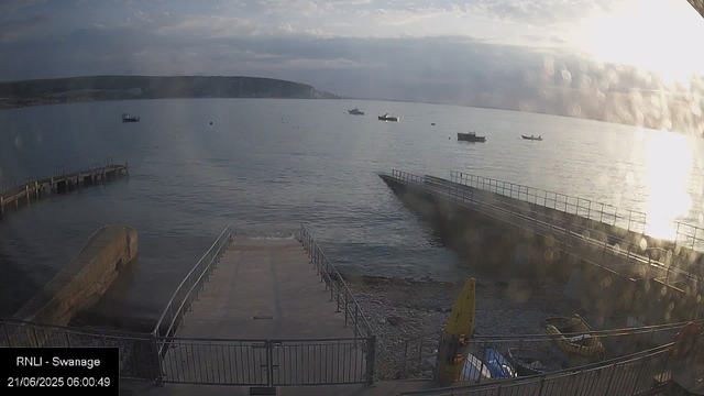 A view of a calm harbor with several boats floating on the water, a small pier extending into the bay, and a walkway leading to the water. The sky is partly cloudy with the sun reflecting off the surface of the water, and a distant landmass is visible on the horizon.