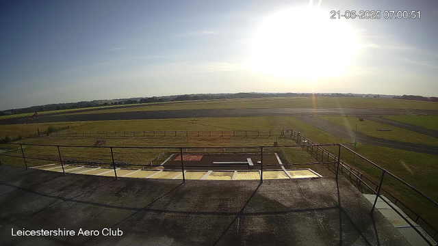 An outdoor scene at the Leicester Aero Club showing a clear sky with the sun shining brightly near the horizon. There are grassy fields and a runway stretching into the distance. The image appears to be taken from a high vantage point, possibly from a building or tower, with a railing visible in the foreground. The timestamp indicates the photo was taken on June 21, 2025, at 07:00:51.