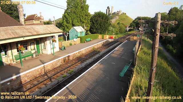 A small railway station platform with a stone building on the left, decorated with hanging flower baskets. There are green benches and a green shelter on the platform. Train tracks run alongside the platform, with a row of wooden utility poles on the right. In the background, a lush green hillside with a castle ruins perched on top is visible, along with trees and a blue sky. The scene is brightly lit, indicating early morning sunlight.