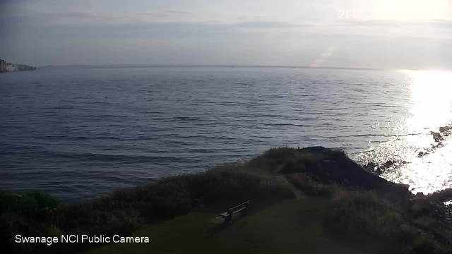A view of the ocean with gentle waves, a rocky shoreline on the right, and a grassy area with a bench in the foreground. The sun is low on the horizon, reflecting brightly on the water. There are cliffs visible on the distant left side. The image is from a webcam labeled "Swanage NCI Public Camera."