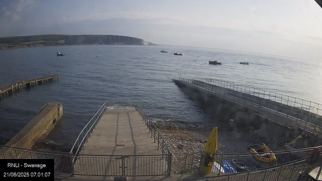 A view of a seafront with calm water and several boats anchored in the distance. A concrete pier extends into the water from the foreground, and there are metal railings along the edges of the pier and the walkway. The background features a hilly coastline under a partly cloudy sky. There are some structures and equipment, including a yellow safety item, on the right side near the walkway, and a small rocky beach area.