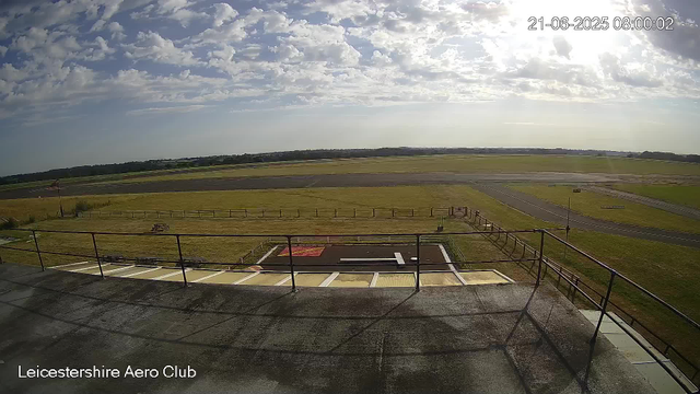 A wide view from a webcam at Leicesterhire Aero Club shows an open sky with scattered clouds during daytime. In the foreground, there is a balcony with metal railings and a small tarmac area with markings, including a red square and a white "H" for helicopter landing. Beyond the balcony, there is a grassy area with a few small structures, a paved taxiway, and open fields stretching towards the horizon. The timestamp in the upper right corner indicates the date as June 21, 2025, at 8:00 am.