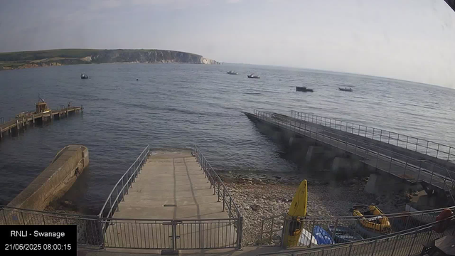 A view from a webcam showing a coastal area with a concrete pier extending into the water. Several small boats are anchored or moving in the sea. There are cliffs in the distance along the shoreline. A yellow caution sign and some life-saving equipment are visible near the bottom right corner of the scene. The weather appears partly cloudy with gentle waves on the water. The timestamp indicates it is morning on June 21, 2025.