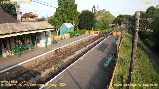 A small train station platform with green benches, a green shed, and flower baskets hanging from the building. Railway tracks run through the center, with a wooden fence and grassy area on one side and a row of telephone poles with wires on the other. In the background, there is a hill with ruins of an old castle or fort. The scene is bright and clear, indicating a sunny morning. The timestamp reads "21-06-2025 08:00:22".