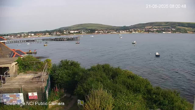 A view of a harbor with several small boats floating on calm water. In the background, there are green hills and buildings along the shoreline. In the foreground, there are trees and some structures near the water's edge. The sky is slightly overcast. The image includes a timestamp showing "21-06-2025 08:00:48".