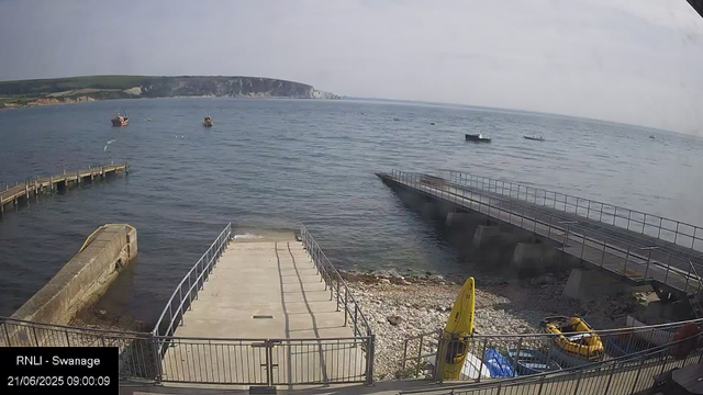 A view of a seaside harbor with a small dock extending into the water. Several small boats are anchored in the water. On the right side, there is a sloped platform leading into the water, with a walkway on top. In the foreground, there are some safety equipment, including yellow life boats, and a yellow caution sign. The background features a rocky coastline with green hills and white cliffs. The timestamp on the image reads 21/06/2025 09:00:09.