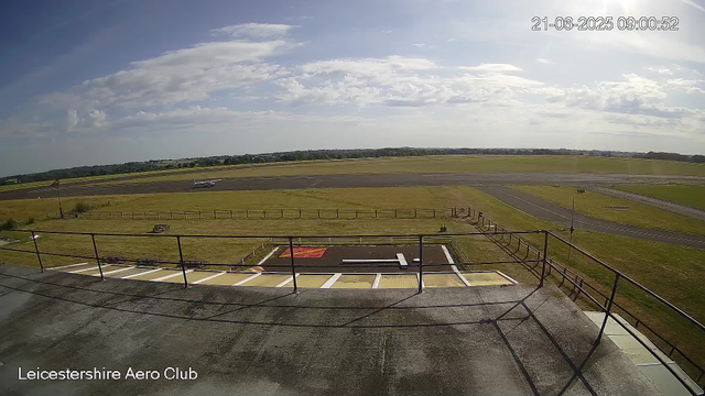 A view from a webcam at Leicester Aero Club showing a runway and grassy airfield under a partly cloudy sky. An aircraft is taxiing in the distance on the runway. The image is timestamped June 21, 2025, at 09:00:52. The foreground includes a balcony with a metal railing, and the club's name is visible in the bottom left corner.