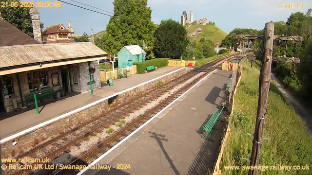A small railway platform with a stone building on the left, decorated with hanging flower baskets, and a green bench in front. The track runs through the center of the image towards a distant hillside with ruins of a castle or fortress on top. There is a green shed behind the platform, surrounded by trees and bushes. On the right, a lamppost casting a shadow on the pavement, and a yellow wooden fence lines the edge of the platform. The sky is mostly clear with some faint clouds. The timestamp indicates it is morning on June 21, 2025.