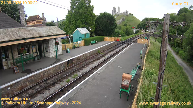 A scenic view of Swanage Railway's old train station platform with a small stone building on the left, decorated with hanging flower baskets. There are green benches, a small turquoise shed, and a yellow picket fence along the platform. The railway tracks run through the center, curving toward a lush green hillside with ruins of a castle on top. To the right, there is a wooden utility pole with several wires, a pathway parallel to the tracks, and grassy overgrowth. In the background, the hillside and castle are visible, and the sky is partly cloudy. The timestamp shows the image was captured on June 21, 2025, at 10:00 AM.
