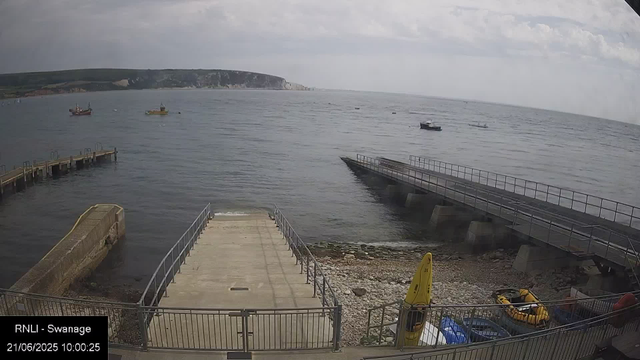 A view of a harbor with concrete piers extending into the water, boats anchored in the distance, a rocky shoreline in the foreground, and cliffs in the background under cloudy skies.