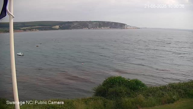 A view of a calm sea with a few small boats floating near the shore. In the background, there are cliffs and a cloudy sky. Green bushes and grass are visible in the foreground, and a flagpole is partially in the frame on the left. The timestamp shows the date as June 21, 2025, at 10:00 AM.
